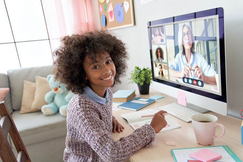 Student of color learning from home sits in front of a computer screen and looks toward the camera with a smile.