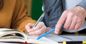 A student and an adult's hands close up. The adult is pointing at the paper with a pen